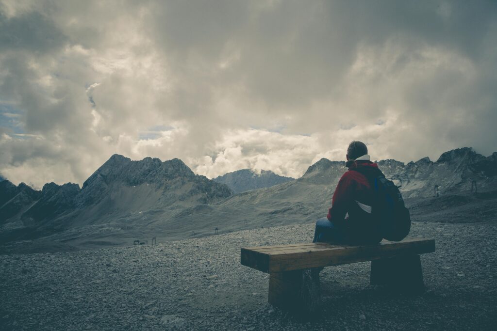 A solitary traveler sits on a bench, gazing at a mountain range under a cloudy sky.