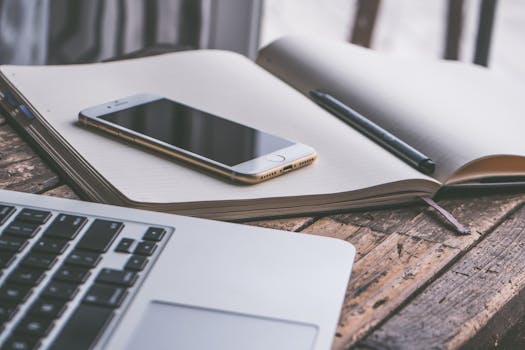Open notebook with a smartphone and laptop on a wooden desk, creating a contemporary workspace vibe.