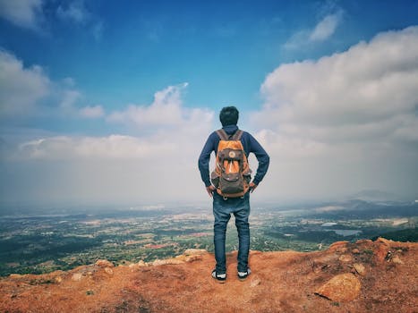 A man stands on a mountain edge appreciating the expansive scenic view below under a bright sky.